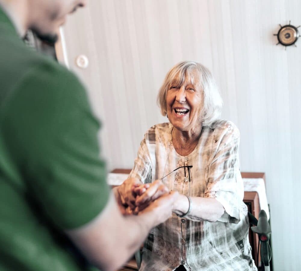 Smiling elderly woman holding hands with a person in a green shirt, sitting indoors. - Home Instead