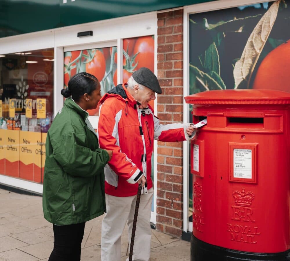 An older male adult with white hair and wearing red jacket and hat mailing a letter with his younger female carer wearing green jacket