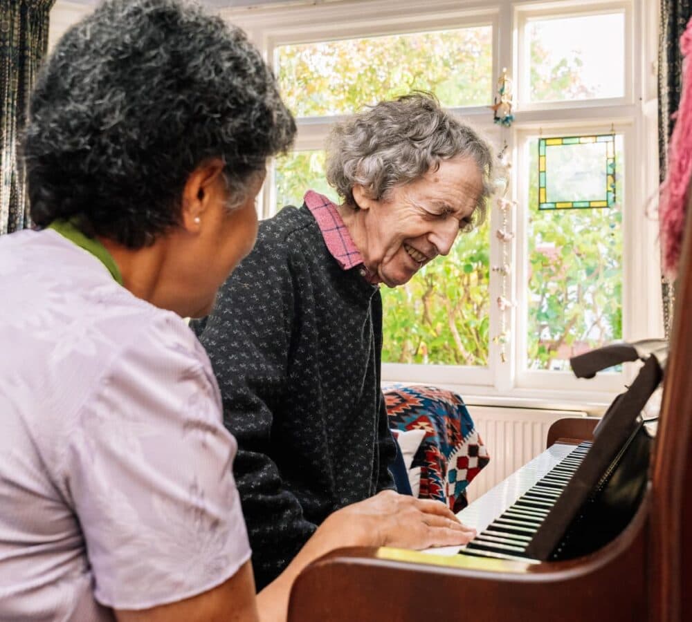 Two elderly women smiling and playing the piano together in a sunlit room. - Home Instead