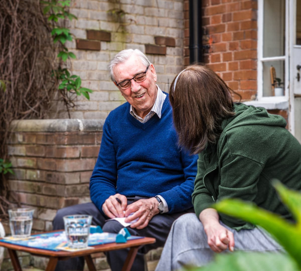 An older male adult with grey hair and wearing eyeglasses and blue sweater playing scrabble while sitting outside the house with his younger female carer