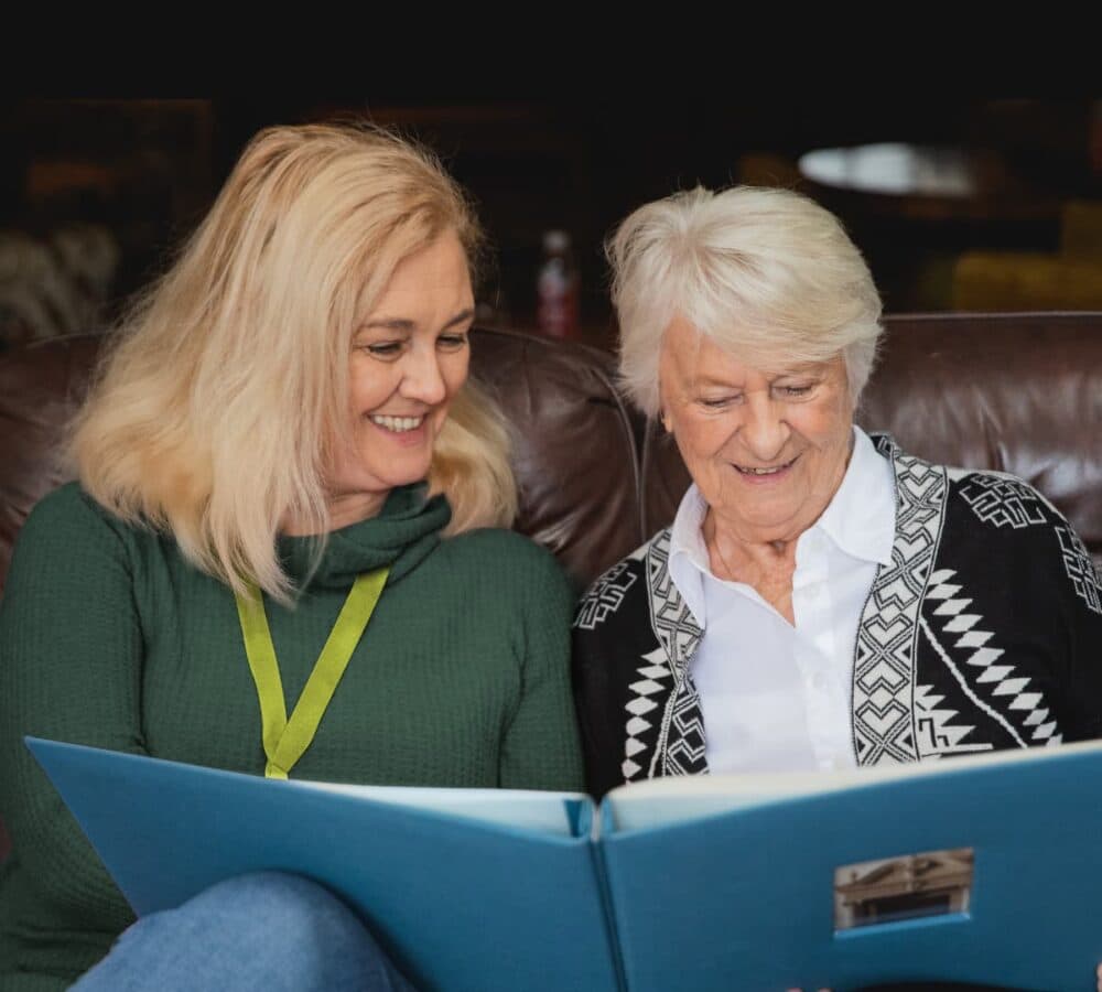 Two women both happy and smiling while looking at a photo album Home Instead