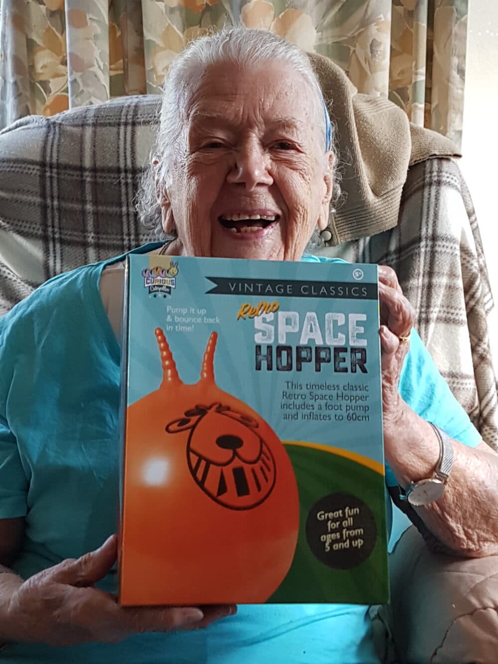 Smiling elderly woman holding a boxed Retro Space Hopper toy while sitting in a chair at home. - Home Instead