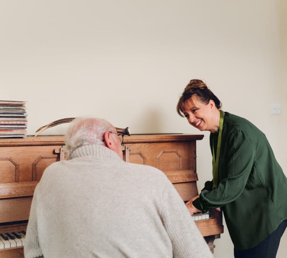 A woman smiles at an elderly man as they play a wooden piano together in a cozy room. - Home Instead