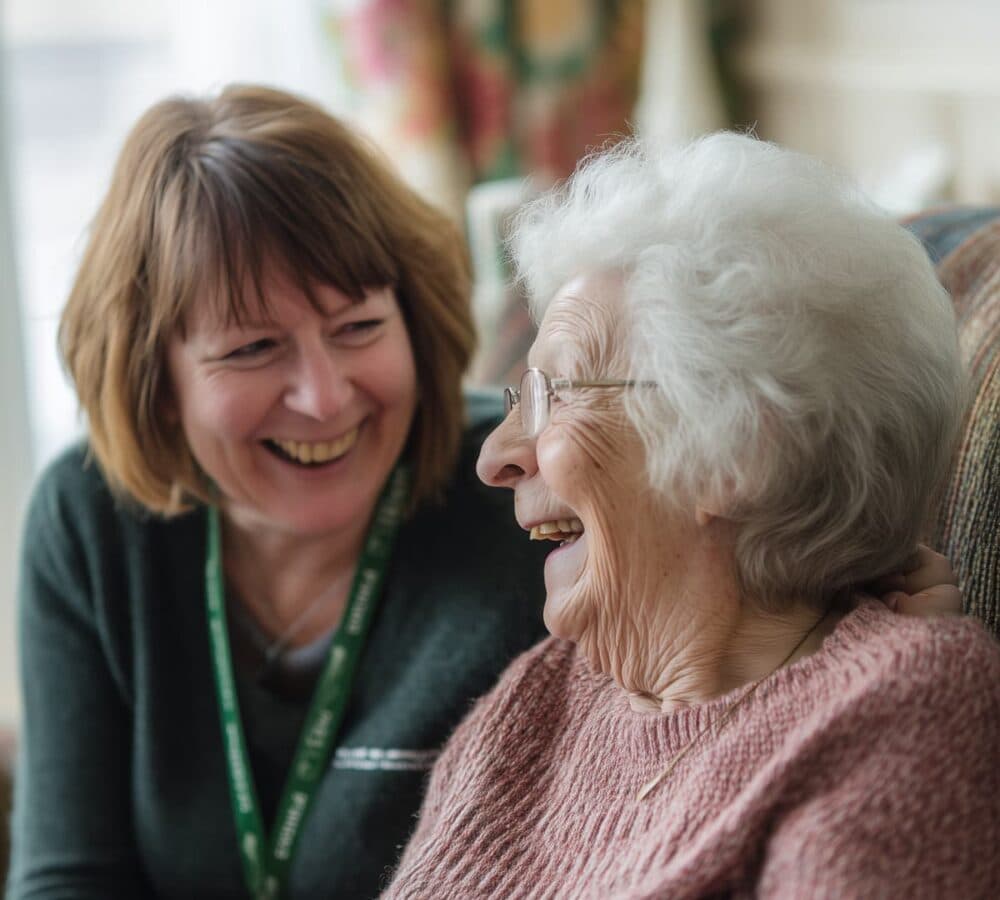 Two women happy and smiling while sitting on a couch inside the house Home Instead