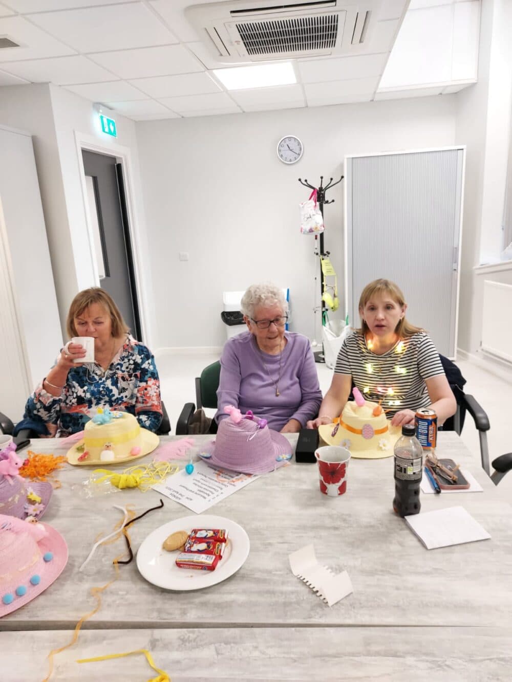 Three women sit at a table with decorated hats, snacks, and drinks, enjoying a casual gathering indoors. - Home Instead