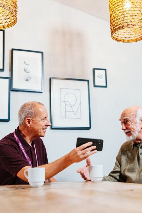 Two older men sit at a table with coffee cups, smiling and looking at a smartphone together. - Home Instead