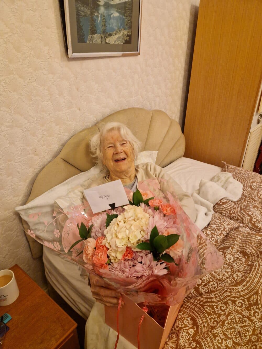 Smiling elderly woman in bed holding a large bouquet of flowers with a card, in a cozy bedroom. - Home Instead