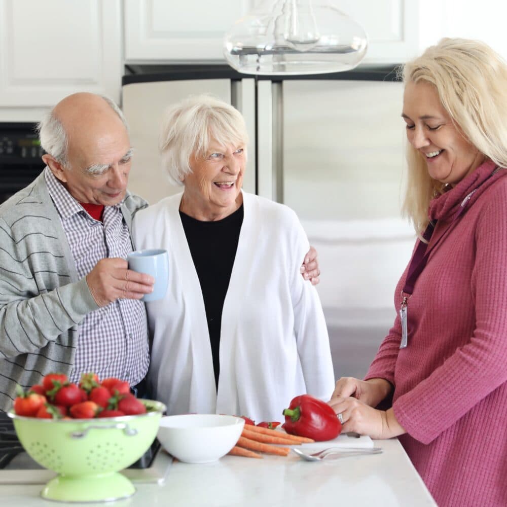 Live-in carer preparing a meal whilst chatting with her male and female live-in care clients in the kitchen.