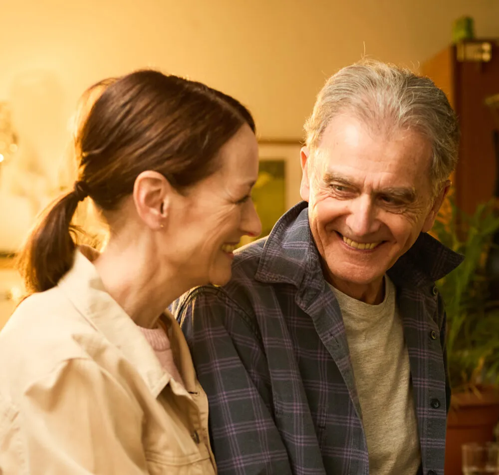 An older man and woman smiling at each other in a warmly lit room. - Home Instead