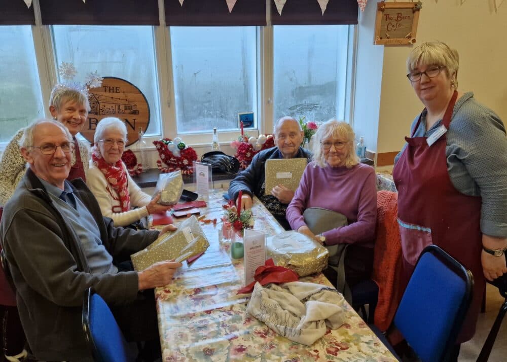 Six elderly people sit around a festive table, holding wrapped gifts and smiling at the camera. - Home Instead