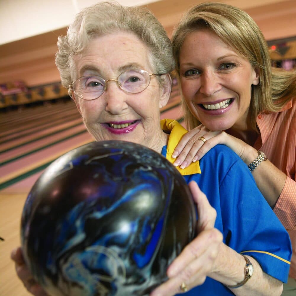 Elderly woman holding a bowling ball in a bowling alley, accompanied by her female carer, both looking as if they are enjoying themselves