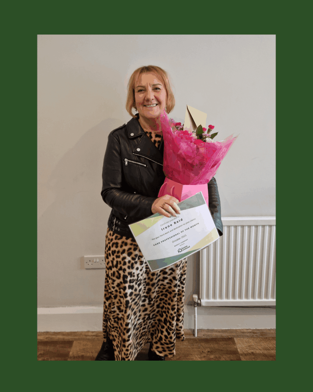 Woman smiling, holding a bouquet of flowers and a certificate, standing against a plain wall. - Home Instead