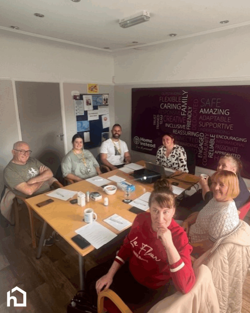 Six people sit around a table in an office, looking up at the camera, with papers and mugs in front of them. - Home Instead