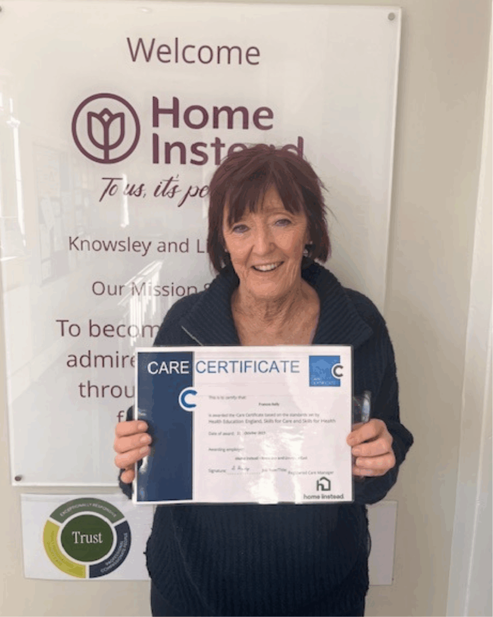 Woman with short hair holding a Care Certificate, standing in front of a Home Instead sign. - Home Instead