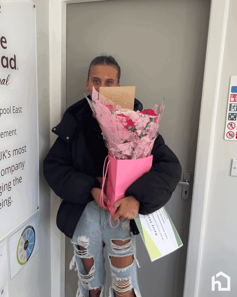 A person in a black jacket holding a pink bouquet and papers, standing in front of a door. - Home Instead