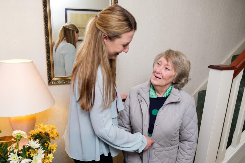 Young female carer helping her elderly female client to put on her coat, standing in the hallway of the client's home.
