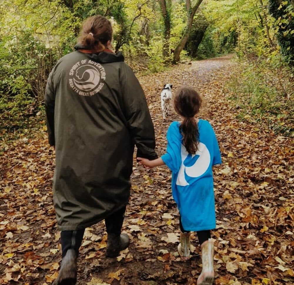 An adult and child hold hands walking a leaf-covered forest path, following a white dog ahead. - Home Instead