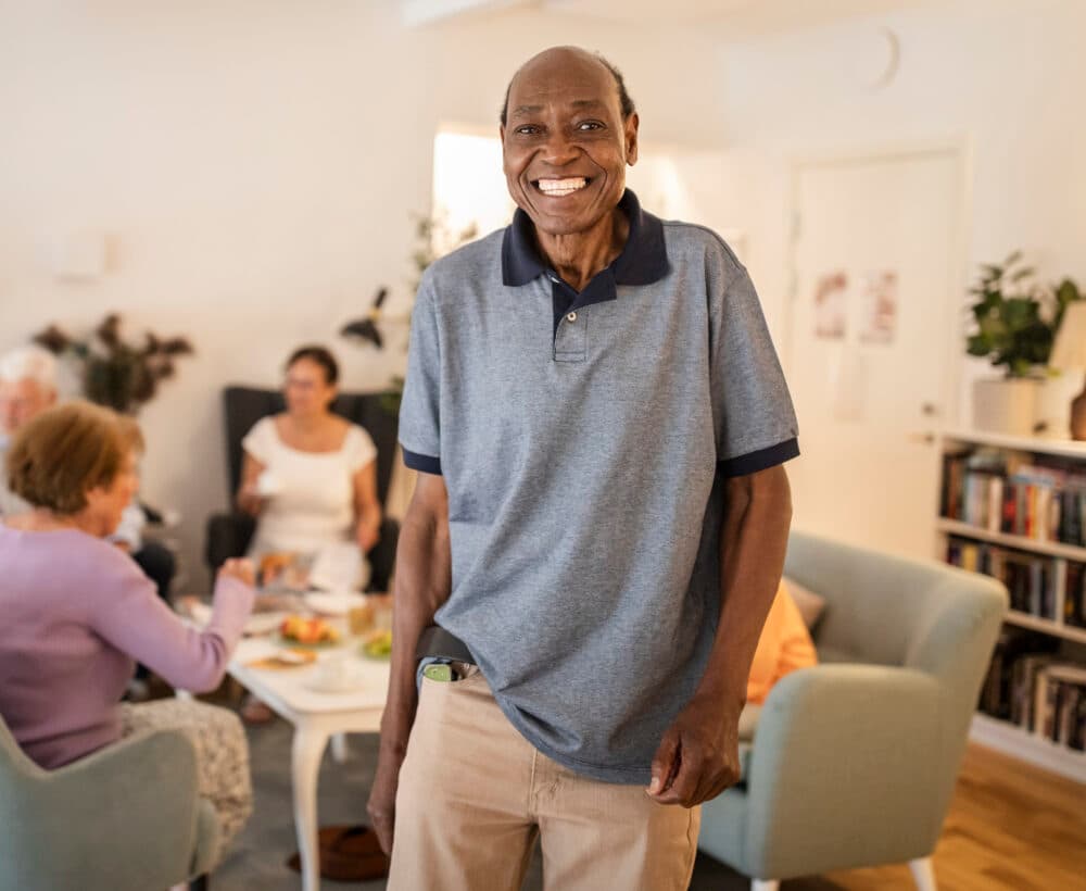 Smiling elderly man stands in a cozy living room with people sitting and talking in the background. - Home Instead