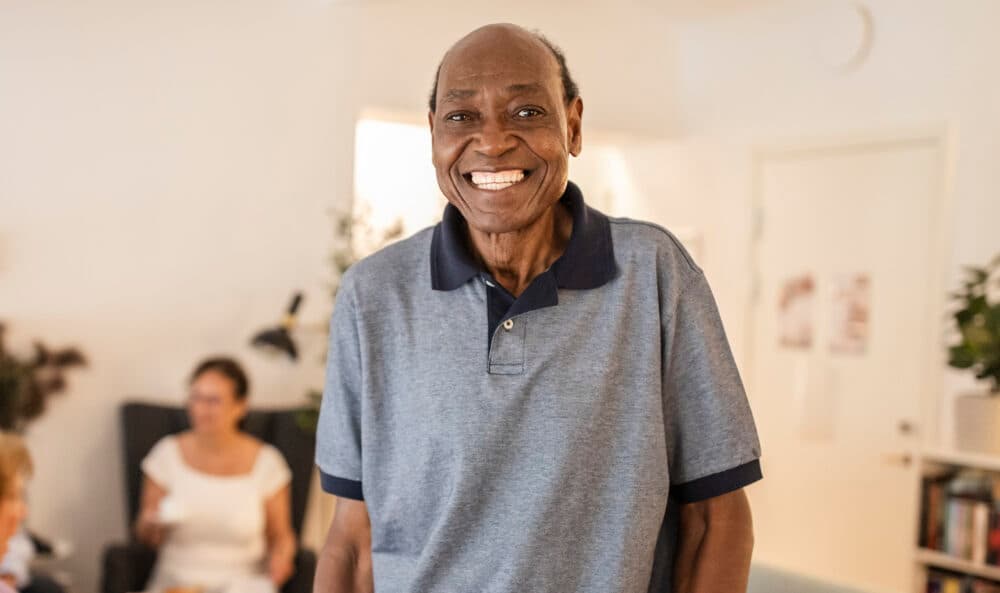 Smiling man in a blue polo shirt standing indoors, with two people seated in the background. - Home Instead