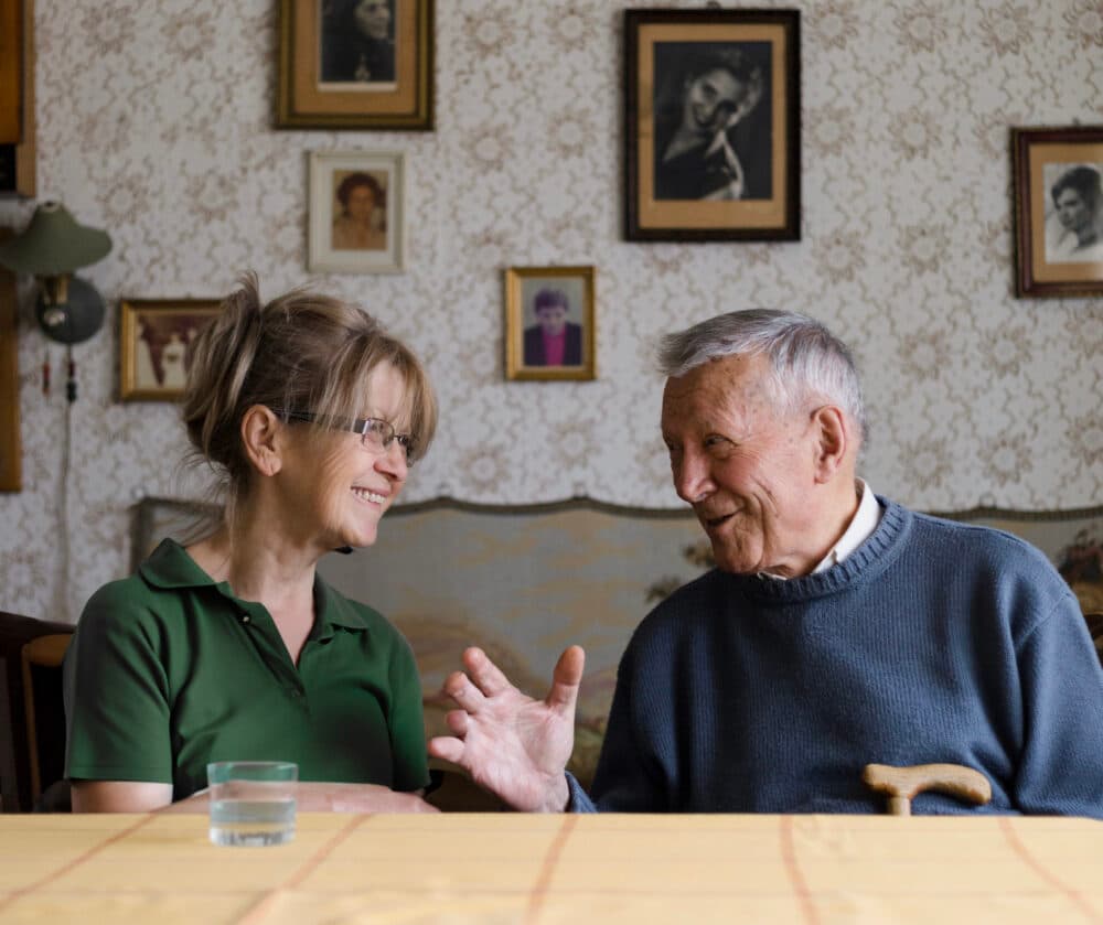 An elderly man and a woman smile and talk at a table, with framed photos on the wall behind them. - Home Instead