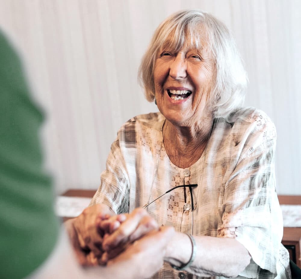 Smiling elderly woman holding hands with another person, sitting indoors and looking happy. - Home Instead