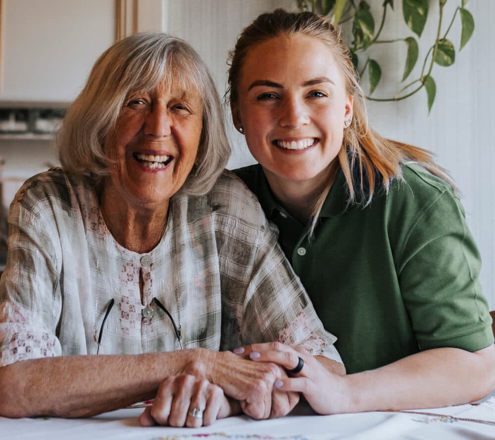 An older woman and a younger woman smiling together at a table, holding hands in a cozy room. - Home Instead