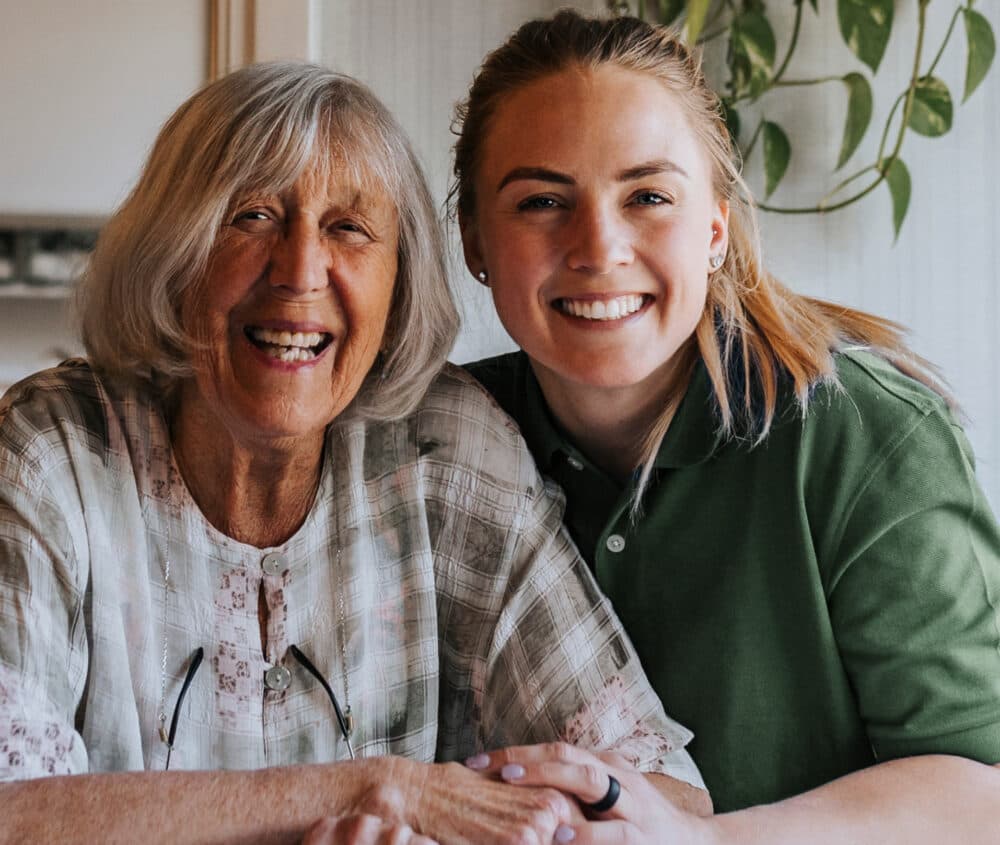 Smiling older woman and younger woman sitting close together, holding hands indoors with a plant in the background. - Home Instead
