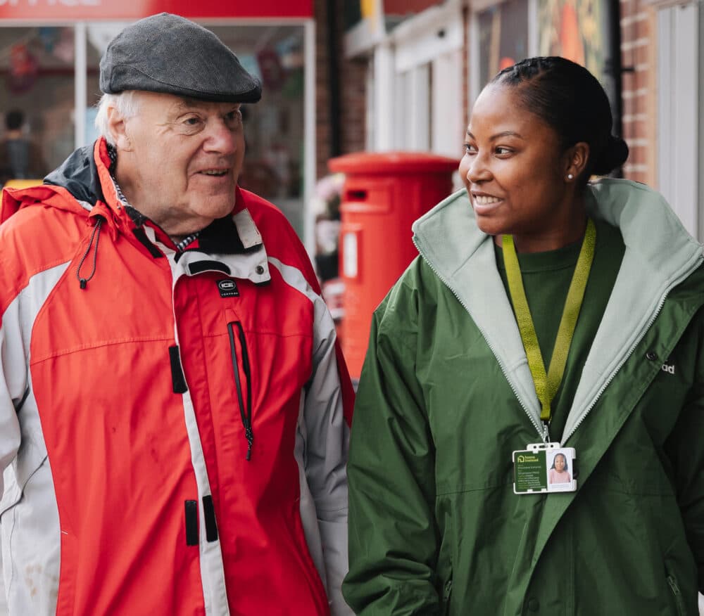 An elderly man and a woman with an ID badge smile while walking together outside. - Home Instead