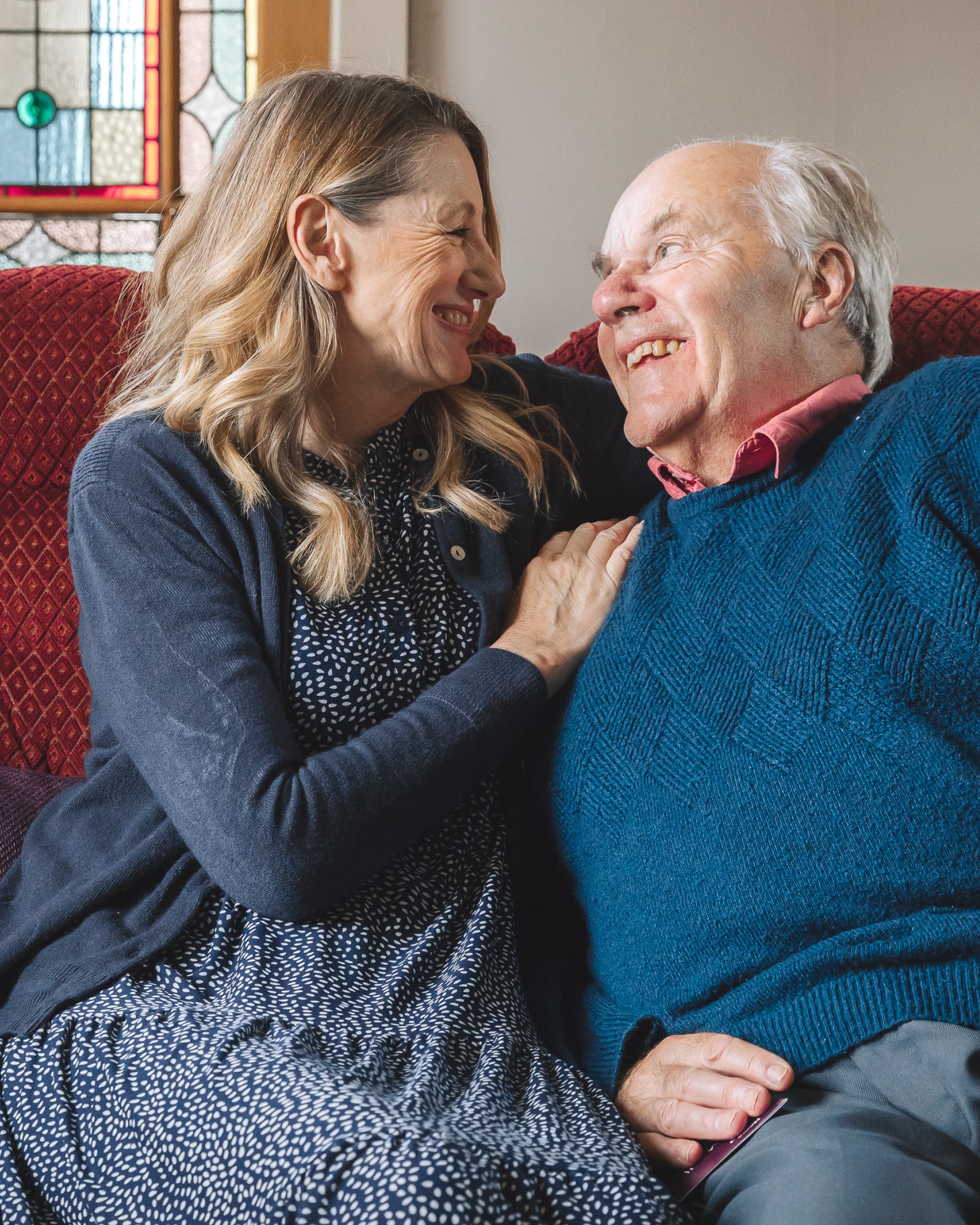 A smiling woman and older man sit closely together on a couch, looking at each other warmly. - Home Instead