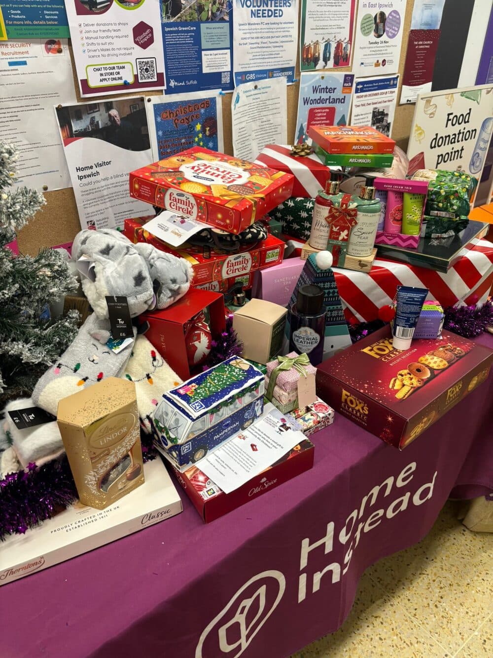Table with Christmas gifts, food, and treats for donation in front of a Home Instead banner and noticeboard. - Home Instead