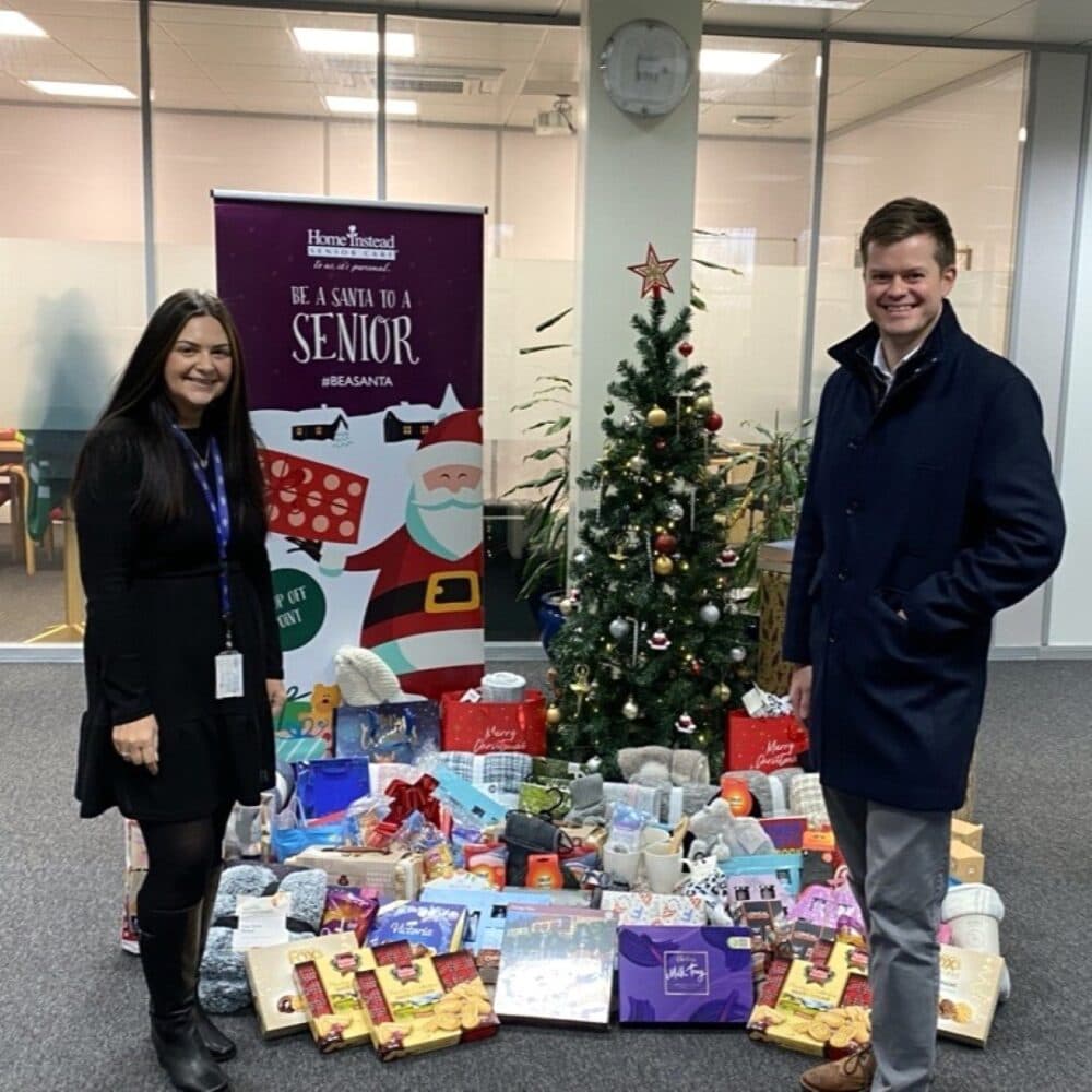 Two people stand by a decorated Christmas tree with gift packages on the floor in an office setting. - Home Instead