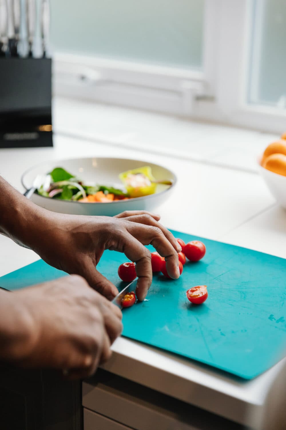 Hands slicing cherry tomatoes on a blue cutting board, with a bowl of salad in the background. - Home Instead