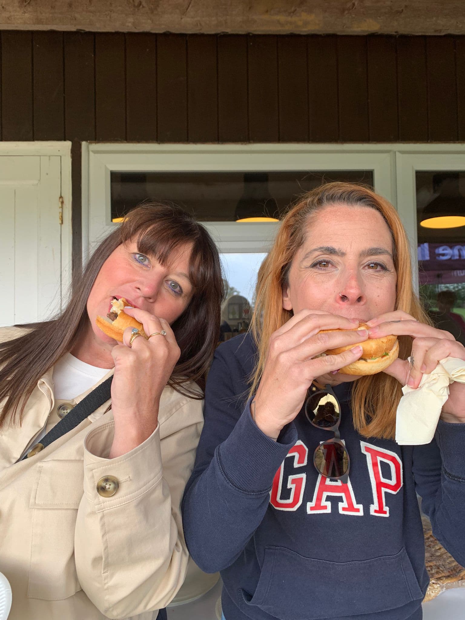 Two women smiling and eating sandwiches outside, one wearing a beige coat, the other a navy GAP sweatshirt. - Home Instead