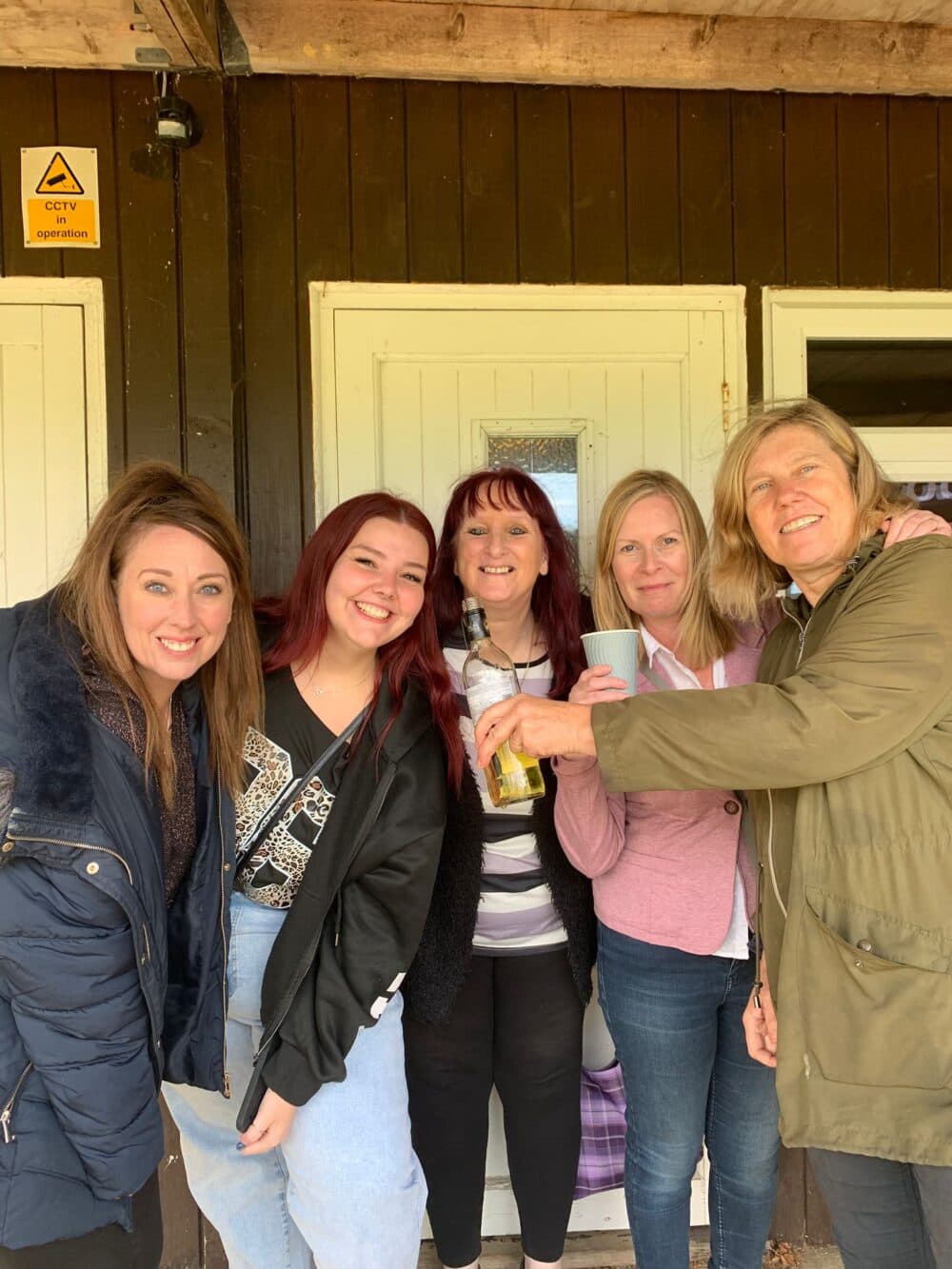 Five smiling women stand together in front of a wooden building, holding drinks and posing for the photo. - Home Instead