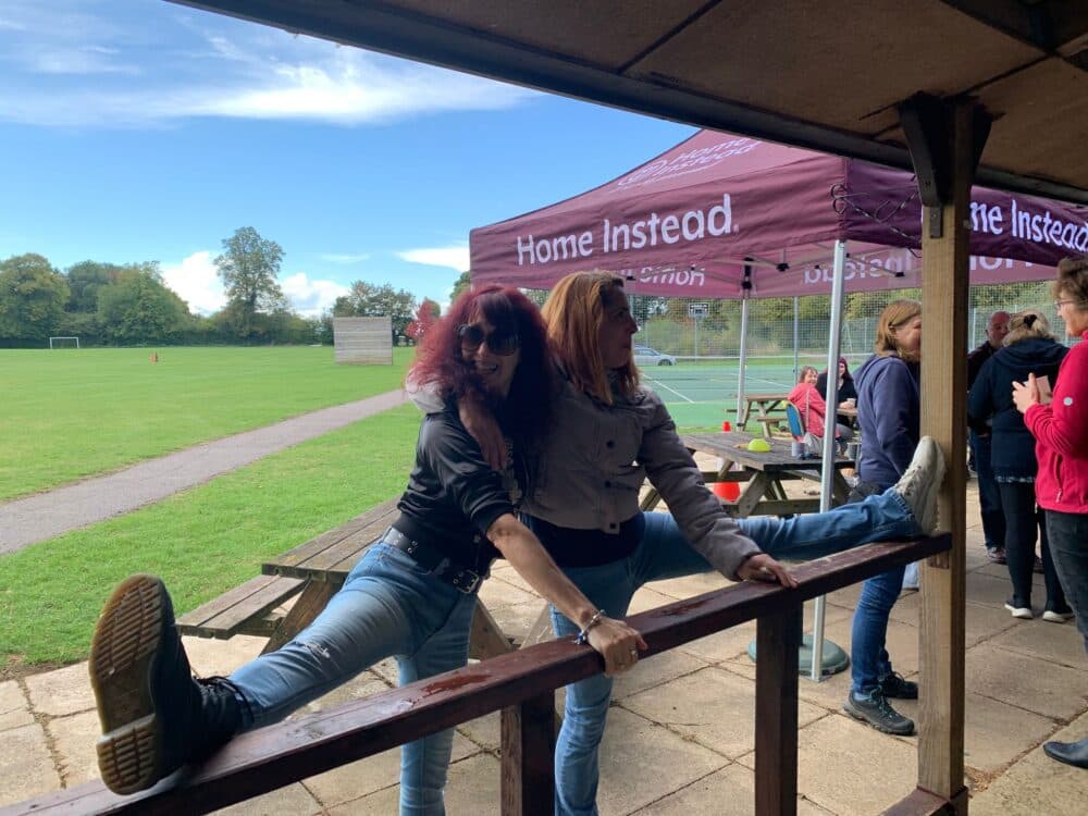 Two women smile while doing splits on a wooden railing at an outdoor event near a sports field and gazebo. - Home Instead