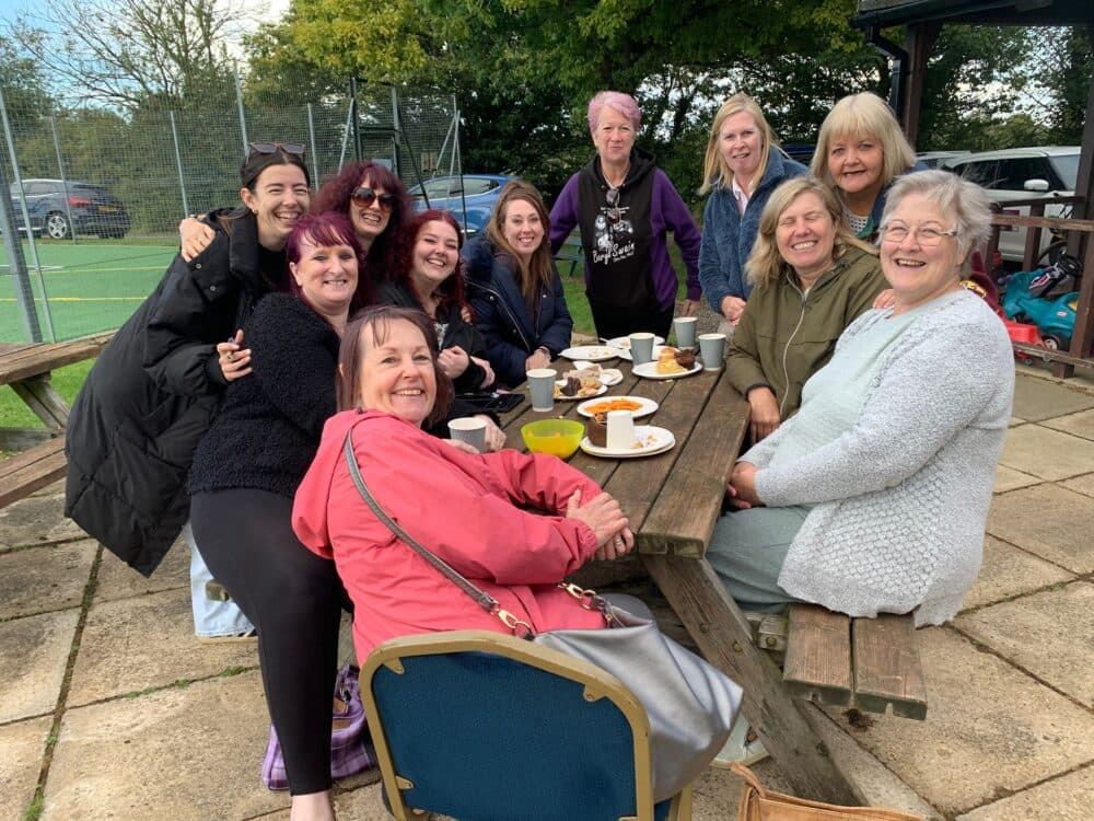 A group of smiling women sit outdoors at a picnic table with drinks and food, enjoying each other’s company. - Home Instead
