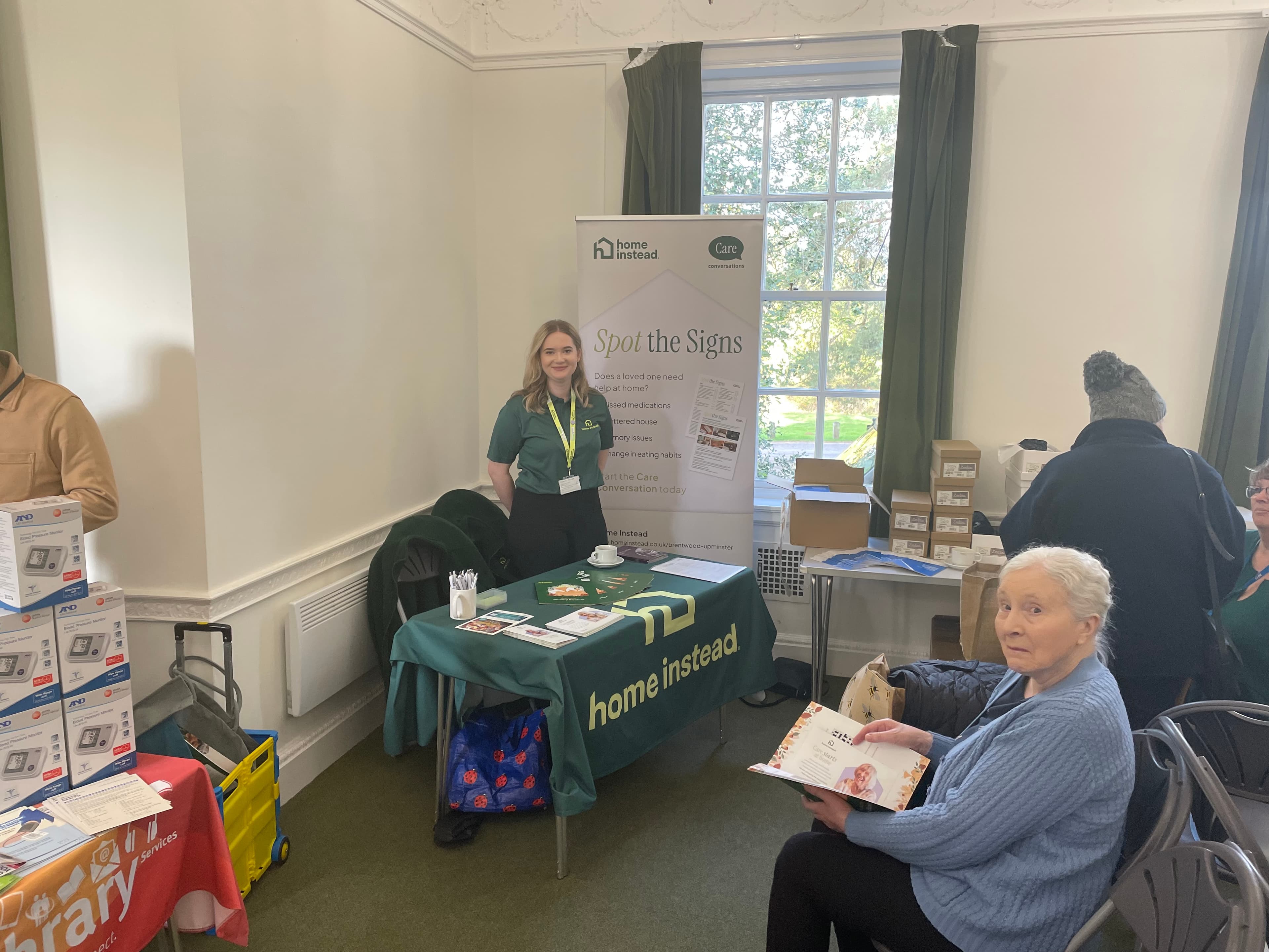 A woman standing in a room with a table with a green tablecloth and a sign. - Home Instead