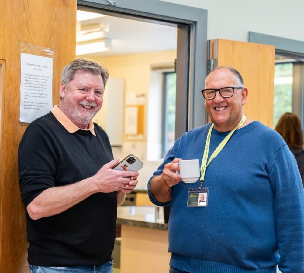 Two men having coffee together both smiling while standing inside the office Home Instead