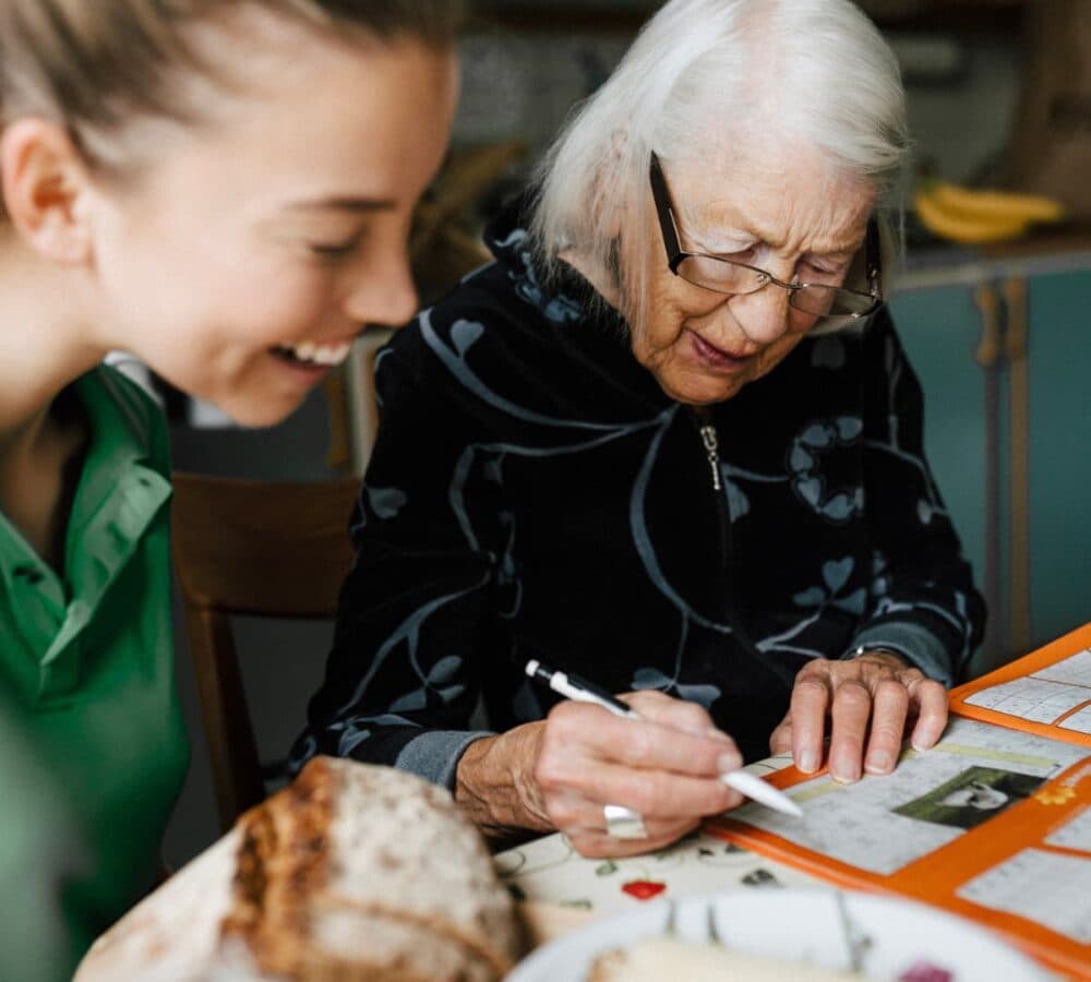 An older female adult with whiteb hair drawing on the table with her younger female carer both happy and smiling