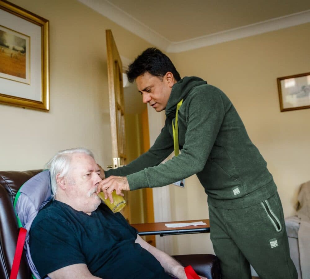 An older male adult with white hair and sitting on a couch while drinking medicine and being helped by his male carer wearing green