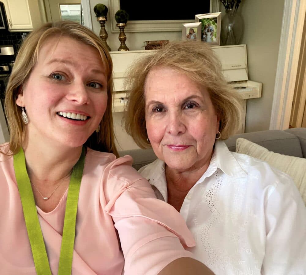 Two women sitting on a couch with blonde hair both smiling and happy while having a photo inside the house Home Instead