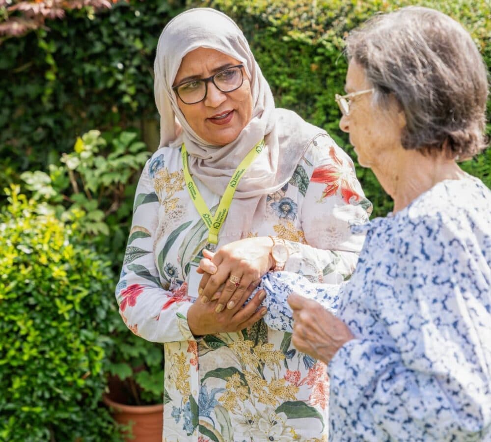 An older female adult with grey hair walking in the garden while being helped by her younger female carer