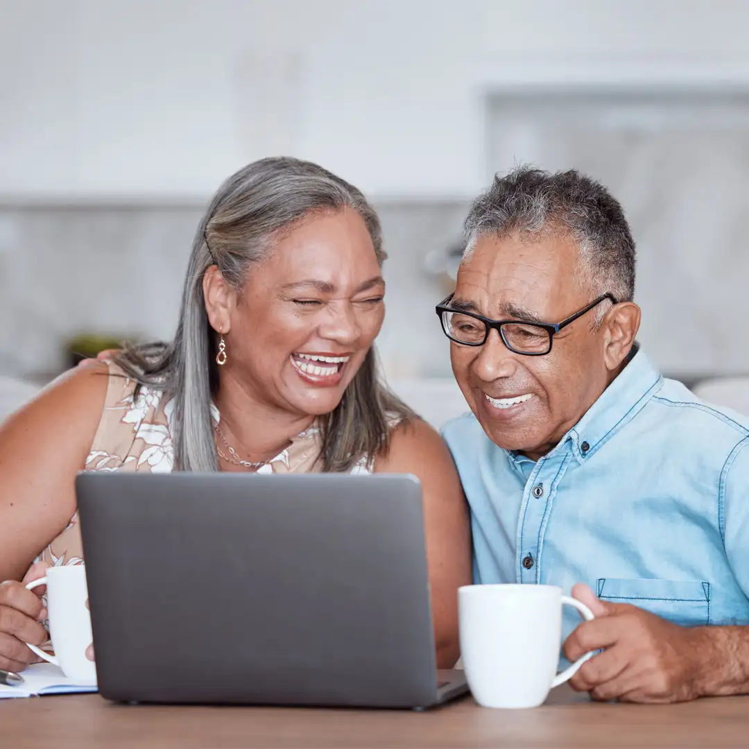 Care pro sat next to an older man supporting him to use his laptop while also enjoying a cup of tea