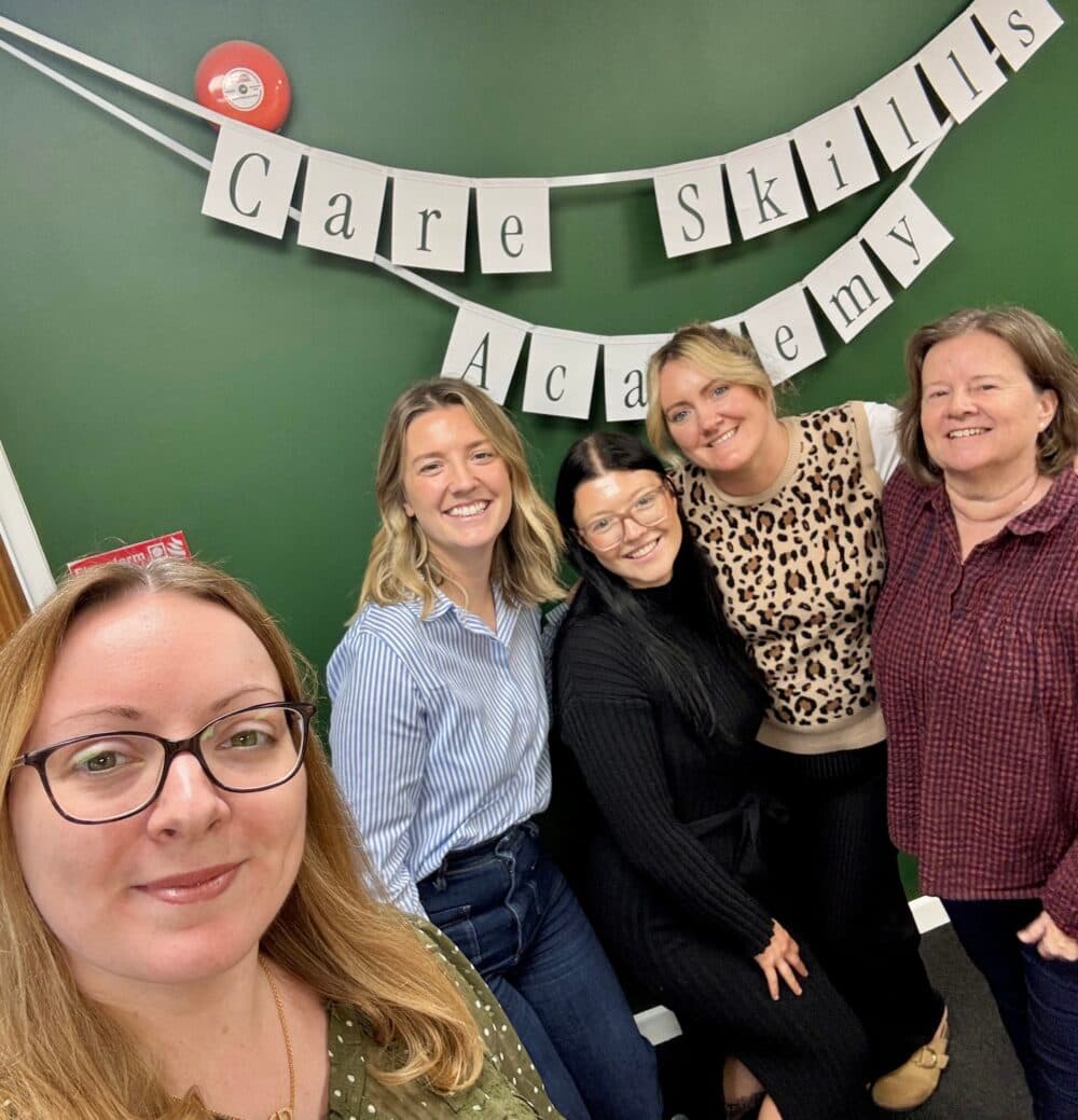 Five women smiling in front of a green wall with a “Care Skills Academy” banner hanging behind them. - Home Instead