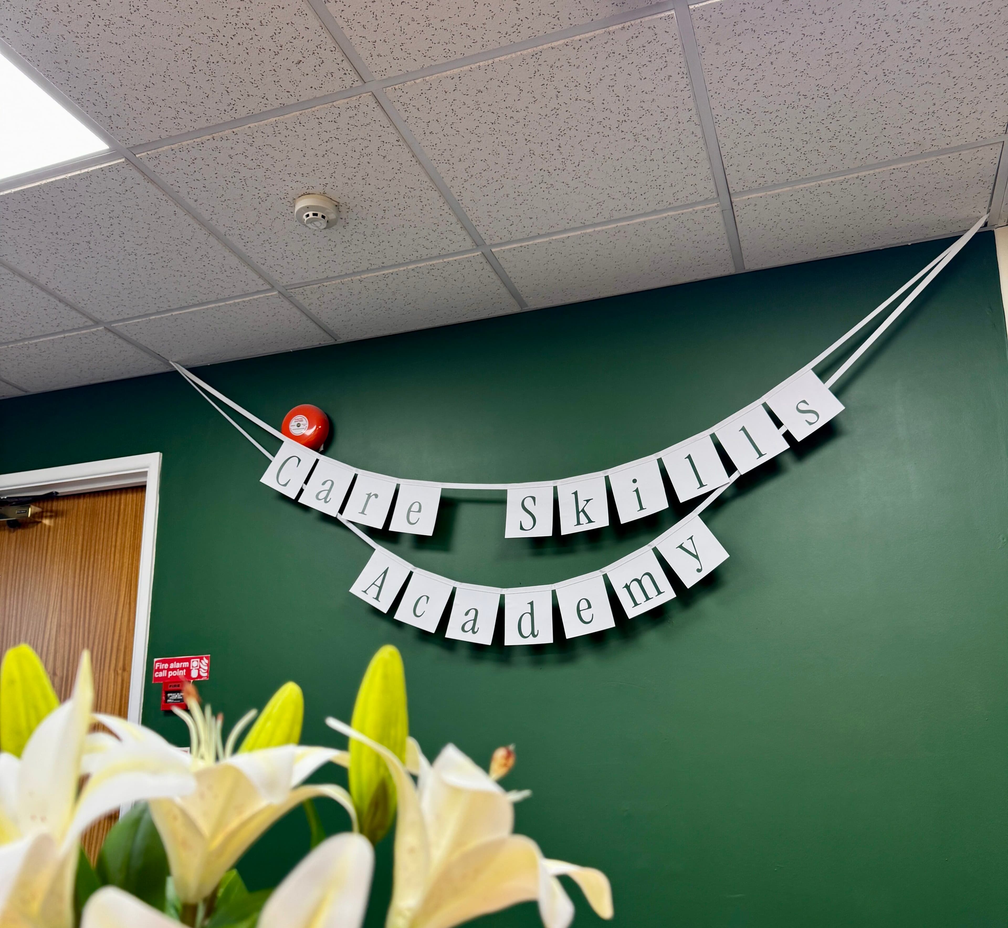 White banners reading "Care Skills Academy" hang on a green wall above white lilies. - Home Instead