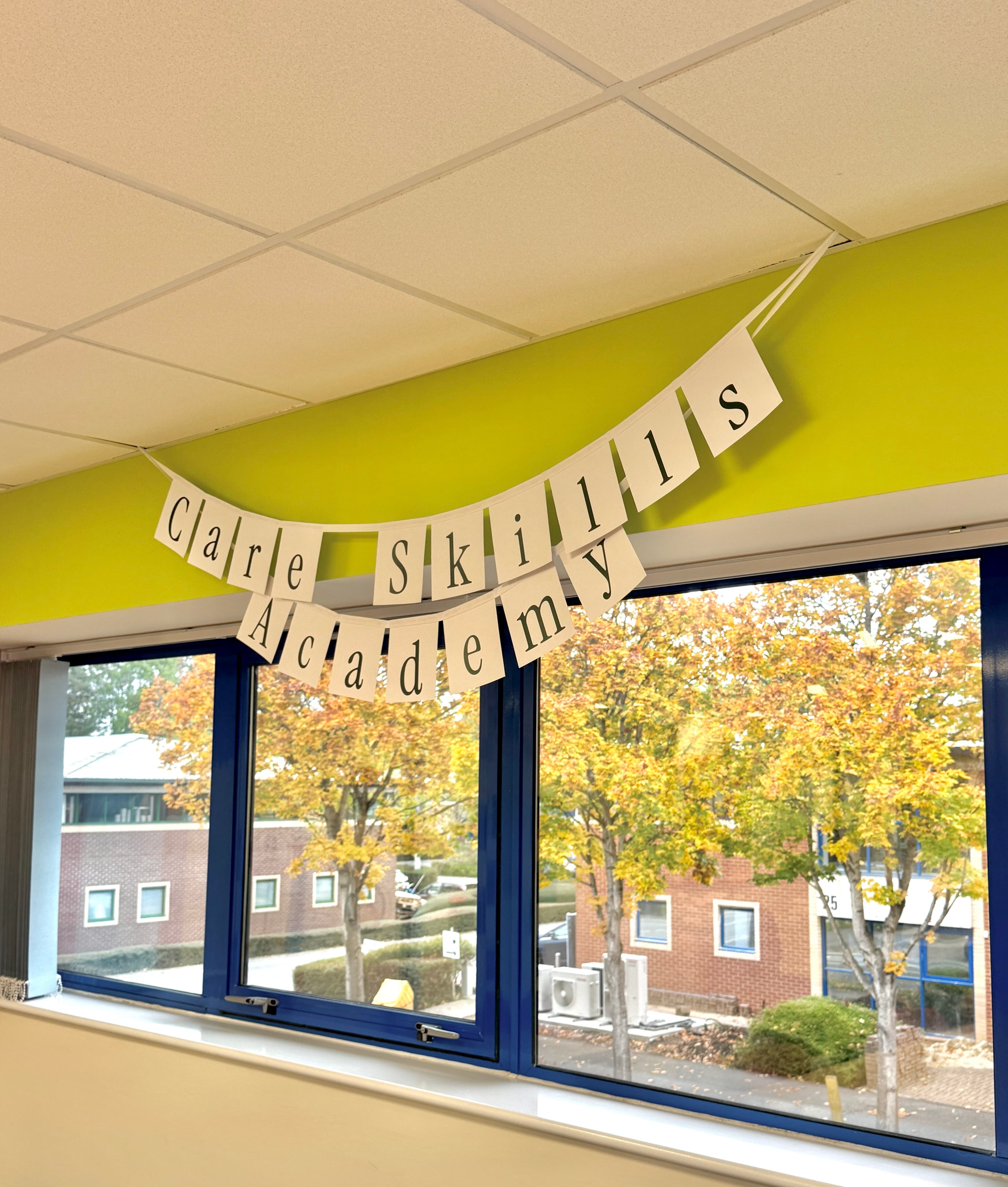 Banner reading "Care Skills Academy" hangs above windows with a view of trees and buildings outside. - Home Instead