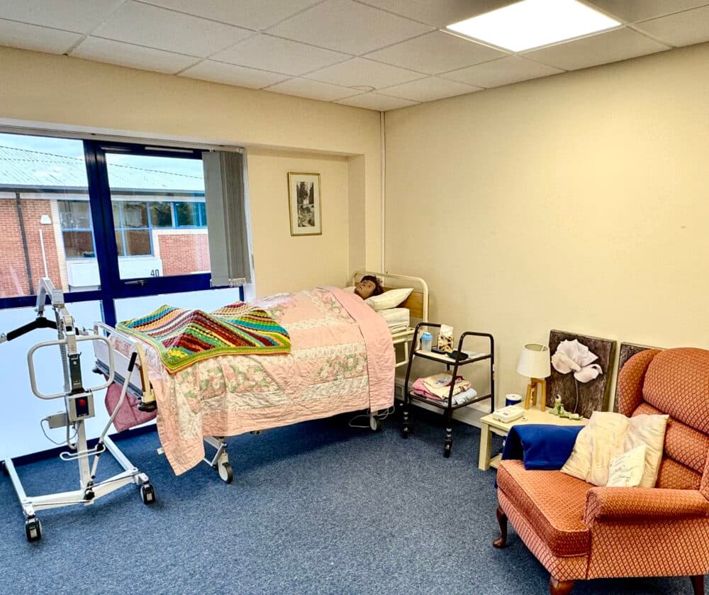 A person lies in a hospital bed in a bright room with medical equipment, a chair, and bedside tables. - Home Instead