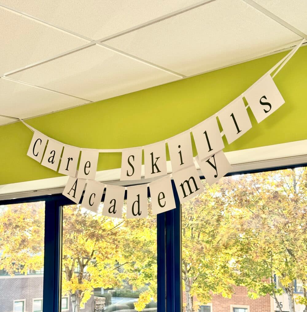 A banner reads "Care Skills Academy" hanging inside near large windows with trees visible outside. - Home Instead