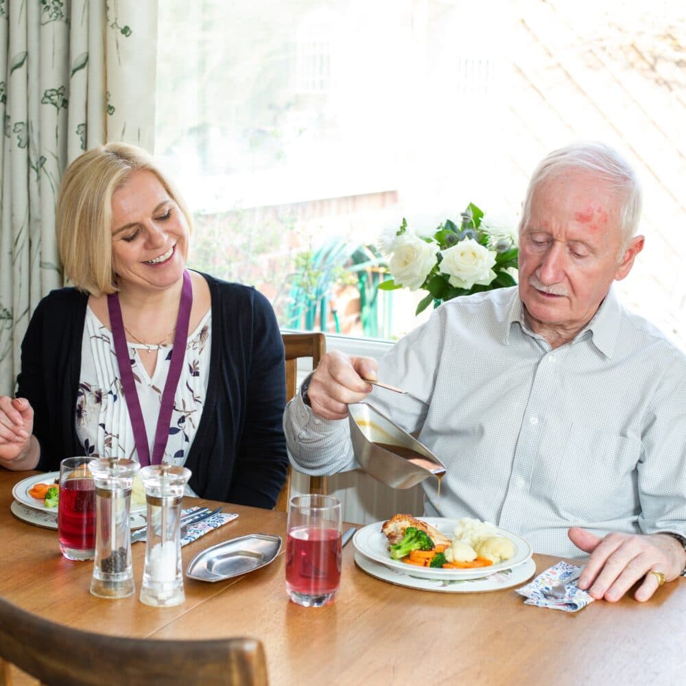 Male live-in client sitting at dining table with his female live-in carer and pouring gravy on his lunch