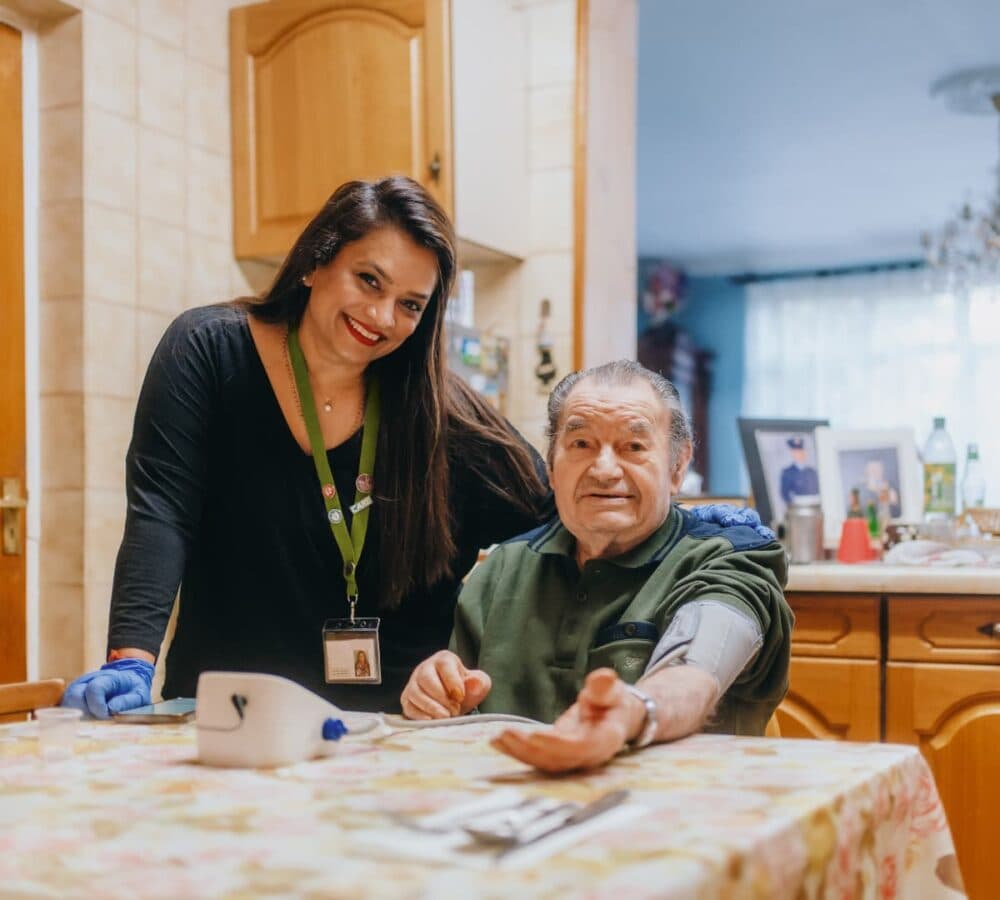 A woman smiles beside an older man having his blood pressure checked at a kitchen table. - Home Instead